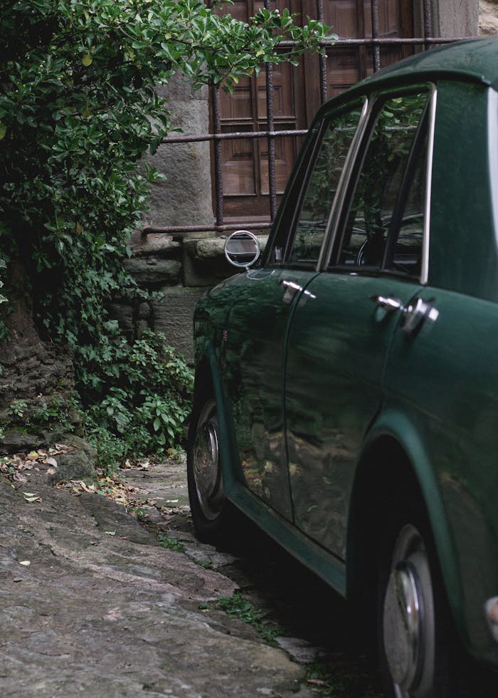 Classic green car parked beside a stone building with greenery.