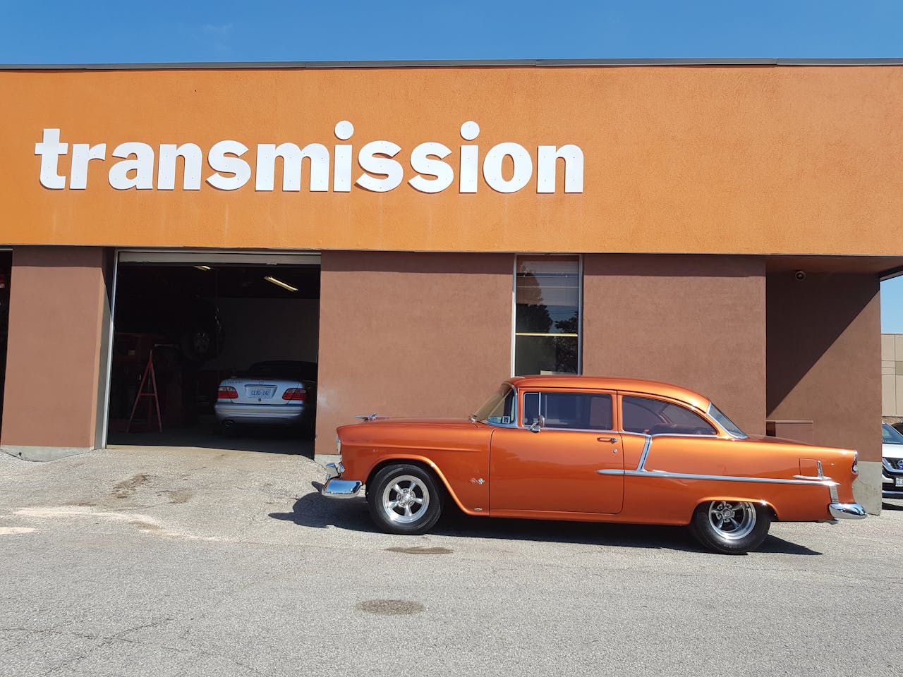 who-we-are A shiny orange classic car parked outside a transmission repair shop on a sunny day.